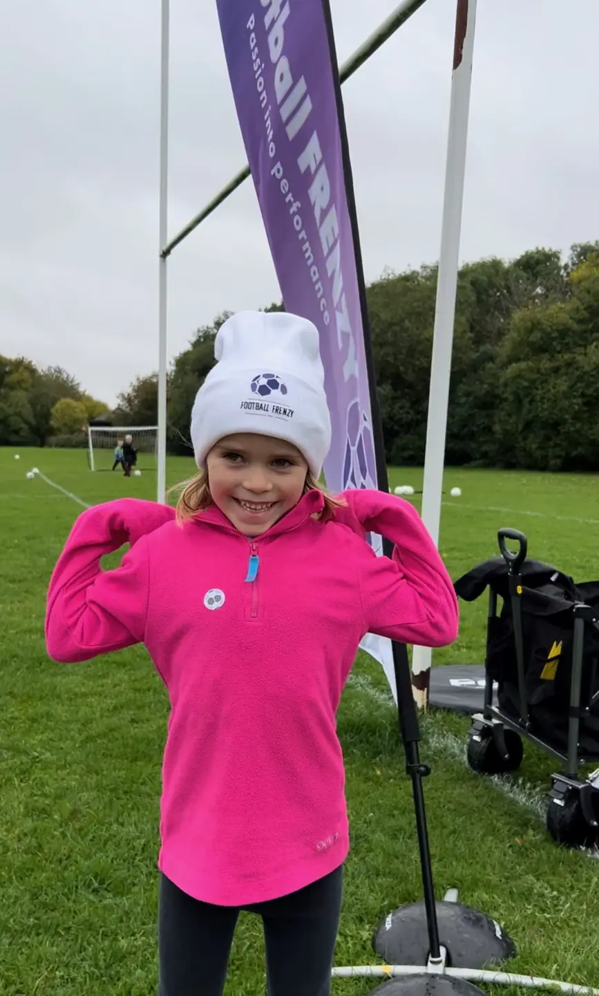 A smiling child in a pink jacket wearing a Football Frenzy hat beside our banner